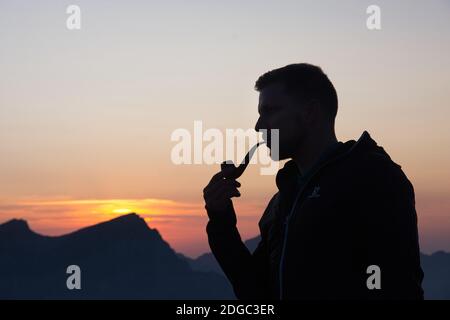 Junger sportlich attraktiver Mann raucht bei Sonnenuntergang am Berg eine Pfeife und genießt die Abendstimmung Stockfoto