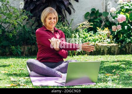 Online Sport Fitness Yoga Training Workout, Senior Frau im Garten ihres Hauses macht Übungen auf Pilates Matte gegenüber Laptop zu Hause. Bus in vi Stockfoto