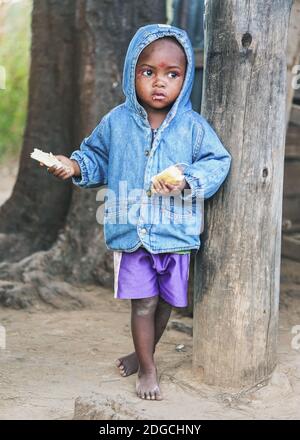 Ranohira, Madagaskar - 29. April 2019: Unbekannter junger madagassischer Junge in blauer Denim-Jacke und Shorts, stehend an Holzpfosten, Brotscheiben essen - Stockfoto