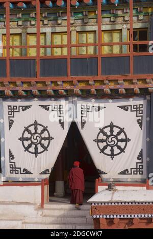 Am frühen Morgen im Hemis Kloster. Mönche betreten den Gebetsraum. Hemis, Ladakh, Jammu und Kaschmir, Indien. Auf dem Stoffschirm am Eingang befindet sich das Dharmachakra - ein typisches Dharma-Rad mit acht Speichen - das den Achtfachen Pfad repräsentiert - das älteste, universelle Symbol für den Buddhismus. Stockfoto