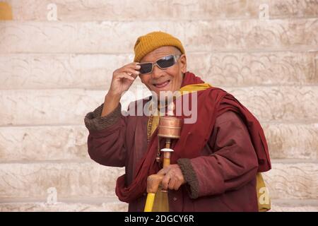 Portrait eines freundlichen, älteren Mönchs mit Sonnenbrille und Gebetrad im Hemis Kloster, Hemis, Ladakh, Jammu und Kashmir, Indien Stockfoto