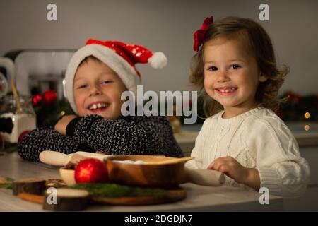 Nettes kleines lächelndes Mädchen 2-4 mit einer roten Schleife und Junge 7-10 in einer Weihnachtsmütze, die Weihnachts Lebkuchenkekse in der Neujahrsküche macht. Stockfoto