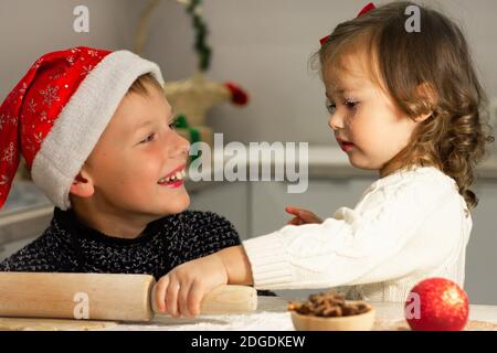 Nettes kleines Mädchen 2-4 mit einer roten Schleife und Junge 7-10 in einer Weihnachtsmütze, die Herstellung von Weihnachts Lebkuchen in der Neujahrsküche.