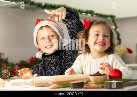 Nettes kleines lächelndes Mädchen 2-4 mit einer roten Schleife und Junge 7-10 in einer Weihnachtsmütze, die Weihnachts Lebkuchenkekse in der Neujahrsküche macht. Stockfoto