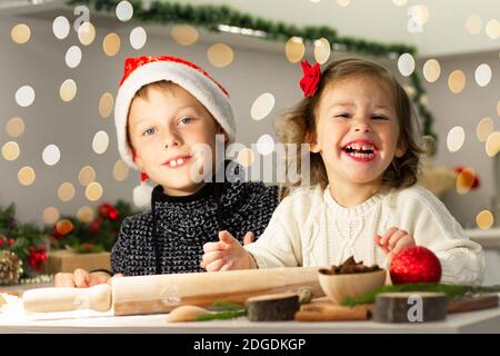 Nettes kleines lächelndes Mädchen 2-4 mit einer roten Schleife und Junge 7-10 in einer Weihnachtsmütze, die Weihnachts Lebkuchenkekse in der Neujahrsküche macht. Stockfoto