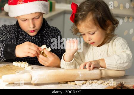 Nettes kleines Mädchen 2-4 mit einer roten Schleife und Junge 7-10 in einer Weihnachtsmütze, die Herstellung von Weihnachts Lebkuchen in der Neujahrsküche. Stockfoto