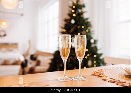 Zwei vergoldete Gläser für Getränke auf dem Tisch, heller gemütlicher Scandi-Raum mit großem Weihnachtsbaum und Lichtern im Hintergrund. Selektiver Fokus Stockfoto