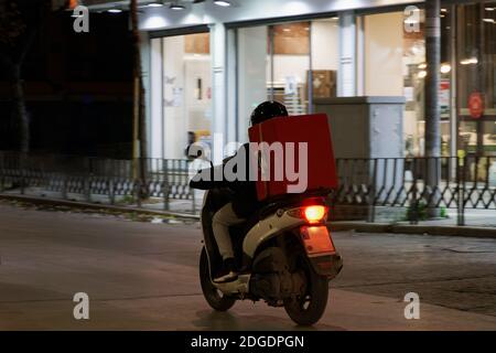 Zustellkurier auf unbranded Motorrad Beschleunigung auf City Road in der Nacht. Beleuchtete Ansicht des Mannes mit Helm Reiten Motorrad zu liefern Essen zum Mitnehmen auf einer roten Box in Thessaloniki, Griechenland enthalten. Stockfoto