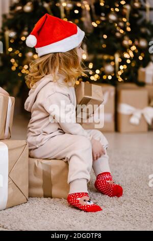 Mädchen in Santa Hut mit Weihnachtsgeschenk auf Weihnachtsbaum Hintergrund. Kind mit Weihnachtsgeschenk zu Hause. Dekoriertes Haus für Winterferien. Weichfokus Stockfoto