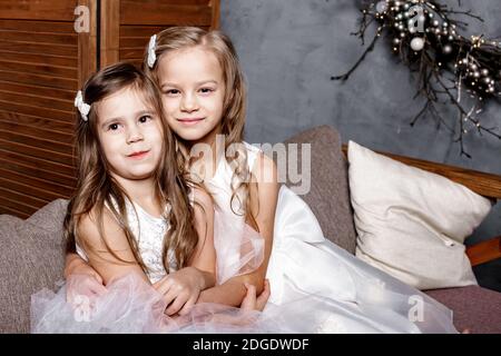 Zwei niedliche Schwestern in weißen Kleidern sitzen auf dem Sofa dicht beieinander und spielen zu Hause. Kleine Schwester umarmt ihre kleinste Schwester. Familie mit Chil Stockfoto