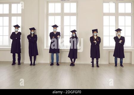 Studenten in Graduierungsroben und Gesichtsmasken stehen im Universitätszimmer, halten soziale Distanz Stockfoto