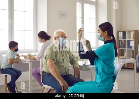 Älterer Mann, der am Tisch im Krankenhaus sitzt und zuschaut Krankenschwester, die Grippeschutzimpfung vorbereitet Stockfoto