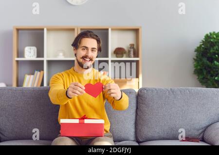 Glücklicher junger Mann mit Geschenk auf seinem Schoß hält Valentine Karte und lächelnd an der Kamera Stockfoto