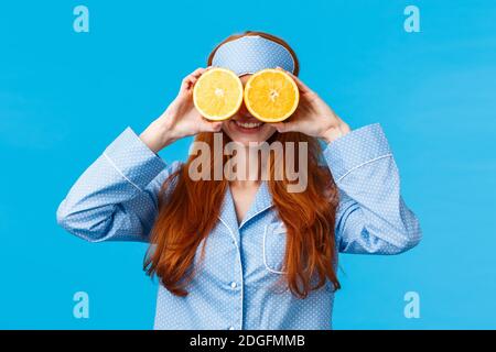 Sorglos hübsche und nette Rotschopf Frau in Nachtwäsche, Schlafmaske, hält zwei Scheiben Orange über den Augen und lächelt, Narr herum Stockfoto