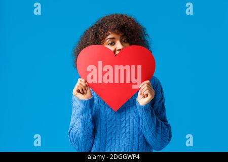 Süßes und liebliches afroamerikanisches Mädchen, mit Afro-Frisur, in Pullover, versteckt Gesicht hinter großen roten Herzen und jubelnd joyfu Stockfoto