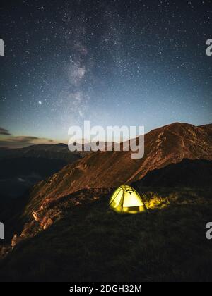 Camping unter Sternenhimmel und Milchstraße in großer Höhe auf den Karpaten. Beleuchtetes Zelt im Vordergrund und majestätischer Berggipfel Stockfoto