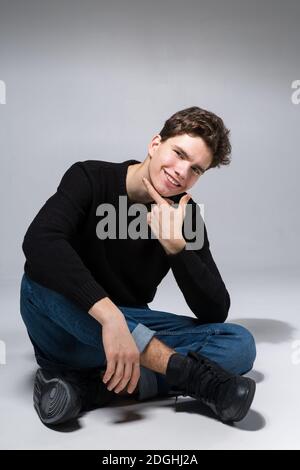 Stilvoller Kerl posiert im Studio sitzt auf dem Boden in Denim-Hosen und schwarzen Sweatshirt. Männlich Student Porträt voll auf weißem Hintergrund Stockfoto