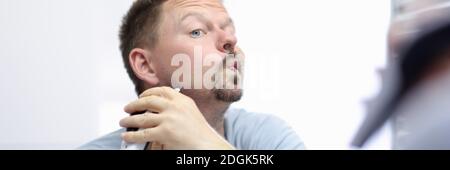 Der junge Mann schaut in den Spiegel und rasiert mit der Maschine Stockfoto