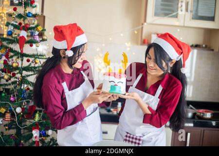 Zwei Glücklich Indischen Mädchen Tragen Schürze Und Weihnachtsmütze Holding Weihnachtskuchen, Feier Während Pandemie. Feiertage und Festtage. Stockfoto