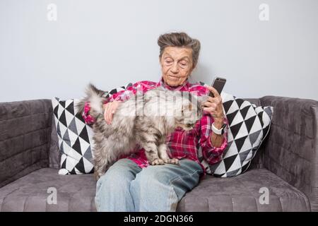 Sehr alte ältere kaukasische Großmutter mit grauen Haaren und tief Falten sitzen zu Hause auf Sofa in Jeans und Hemd mit Grau, flauschig Stockfoto