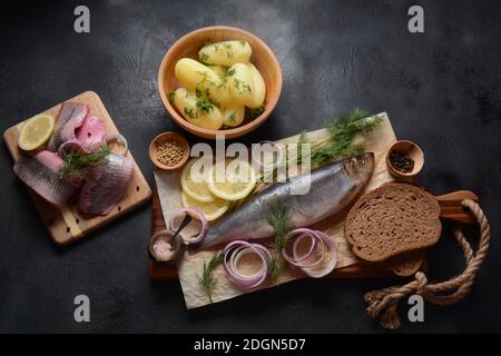 Heringsfisch auf Holzbrett mit Pfeffer, Kräutern, roter Zwiebel und Zitrone auf schwarzem Hintergrund. Draufsicht mit Kopierraum mit Kartoffelscheiben und Roggenfarbe Stockfoto