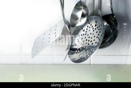 Ein Skimmer, zwei Gabeln und zwei Metallpfannen hängen an der gefliesten Küchenwand. Küchenutensilien aus Metall Stockfoto