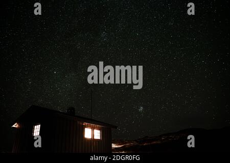 Kleine Hütte in den Bergen mit Licht aus den Fenstern und einem sternenklaren Nachthimmel im Hintergrund. Zeitraffervideo. Stockfoto