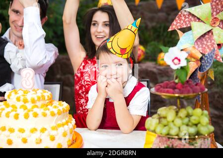 Kindergeburtstag Partymädchen sitzt an einem Tisch in der Nähe eines Kuchens in einem festlichen Hut und lächelt Stockfoto