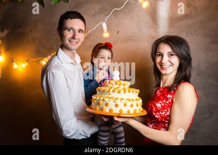 Thema Familienurlaub Kinder Geburtstag und ausblasen Kerzen auf großen Kuchen. Junge Familie von drei Personen stehen und halten Stockfoto