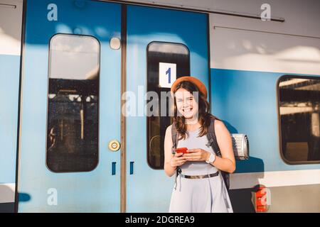 Thema Eisenbahn. Schöne junge Frau mit Rucksack nutzt das Telefon, während sie in der Nähe des Eisenbahnzuges auf dem Bahnsteig steht. Che Stockfoto