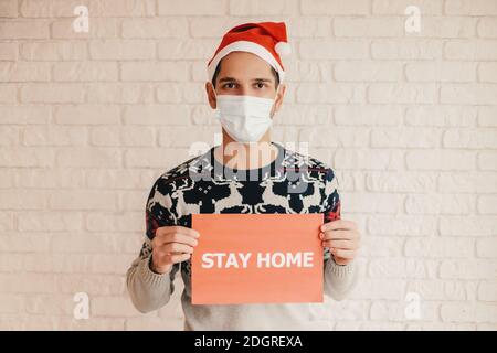 Glücklicher Mann in Santa Hut, medizinische Gesichtsmaske mit zu Hause bleiben Text in den Händen. Junge fröhliche Mann in Weihnachten Pullover halten zu Hause bleiben Banner. Selbstisolierung Stockfoto