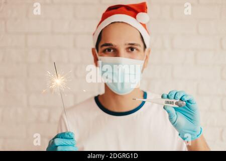 Glücklicher Mann Arzt in Santa Hut, medizinische Gesichtsmaske halten elektronische Thermometer und funkelndes Licht in der Hand. Junger männlicher Chirurg in Schutzmaske, Handschuh Stockfoto