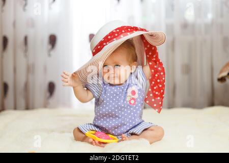 Ein kleines lustiges kaukasisches kleines Mädchen ein Kind sitzt zu Hause auf dem Boden auf einem leichten Teppich. Auf seinem Kopf ist ein großer Sommerhut, die Stockfoto