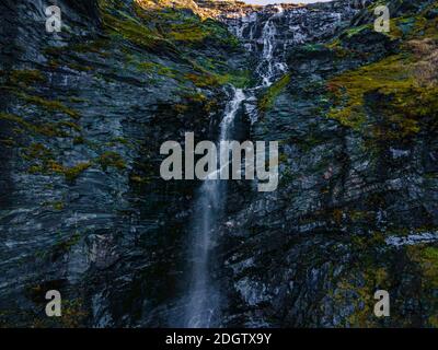 Langer, schmaler Wasserfall, der das Gelände auf einem steilen grünen Berghang durchschneidet. 4K Luftaufnahmen. Stockfoto