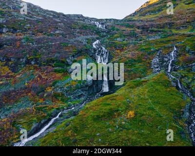 Langer, schmaler Wasserfall, der das Gelände auf einem steilen grünen Berghang durchschneidet. 4K Luftaufnahmen. Stockfoto