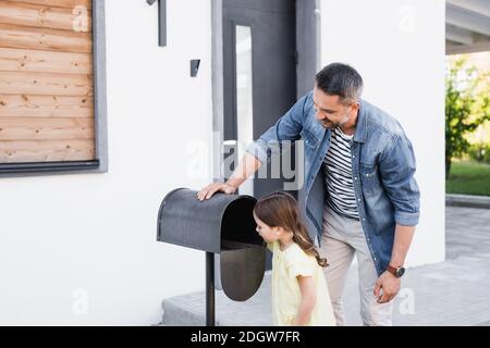 Lächelnder Vater in der Nähe der Tochter stehend und in der Nähe des leeren Briefkastens schauend Haus Stockfoto