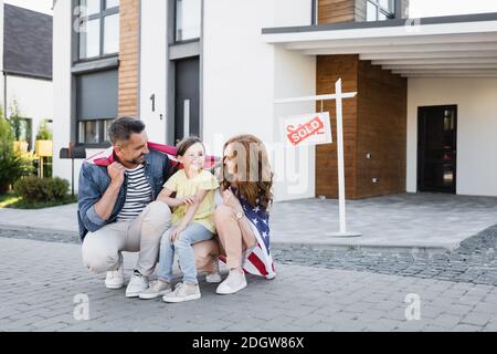 Glückliches Paar mit amerikanischer Flagge, die Tochter beim Hocken ansieht In der Nähe Schild mit verkauft Schriftzug und Haus Stockfoto