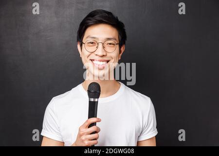Freizeit, Leute und Musik Konzept. Schöner und netter lächelnder asiatischer Mann in weißem T-Shirt, Brille, hält Mikrofon, singt an Stockfoto