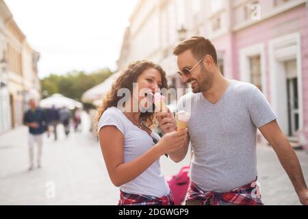 Der junge, gutaussehende Mann gibt seiner Freundin sein Eis aus der Hand auf einer Straße zu essen. Stockfoto