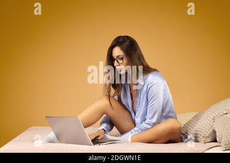 Attraktive junge Frau in blauem Hemd, Brille trägt, sitzt auf ihrem Bett, arbeitet auf ihrem Laptop Stockfoto