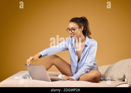 Attraktive junge Frau in blauem Hemd, Brille trägt, sitzt auf ihrem Bett, arbeitet auf ihrem Laptop Stockfoto