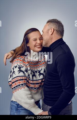 Erwachsener Mann küsst seine glücklich lachende Frau. Liebevolles Paar in warmen Pullovern posiert für Familienportrait. Winterurlaub Fotoshooting Konzept im Studio Stockfoto