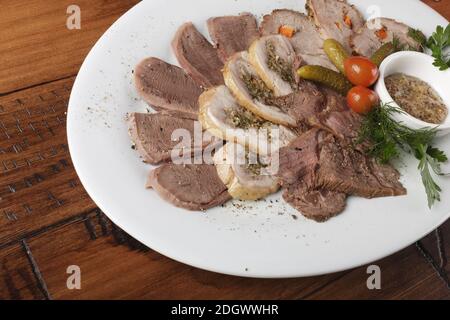 Gebratenes Roastbeef, Hühnerbrötchen, Zunge und Schweinefleisch mit Gemüse auf einem weißen ovalen Teller. Holzhintergrund. Stockfoto