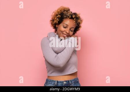 afroamerikanische Frau umarmt sich glücklich und positiv, lächelnd zuversichtlich. Selbstliebe und Selbstfürsorge Stockfoto