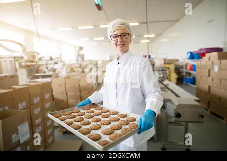 Junge schöne engagierte weibliche Arbeiter in sterilen Tüchern tragen Cookies auf Zinnpfanne innerhalb der gesunden Lebensmittel Produktionslinie. Stockfoto