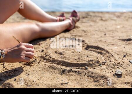 Ein junges Mädchen in einer Weste mit Fernglas sitzt auf dem Sand in der Nähe des Meeres in der Stadt und zeichnet ein Herz mit einem Stock. Stockfoto