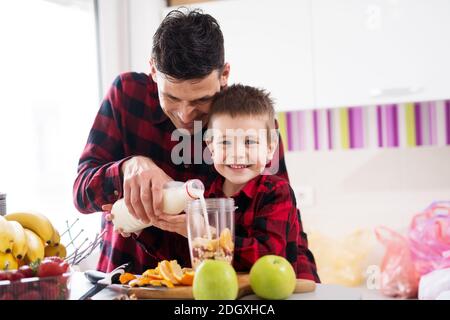 Netter kleiner Junge und sein fürsorglicher Vater gießen Milch in Fruchtmischung in Mixer Schüssel. Stockfoto