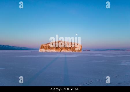 Fantastischer Sonnenuntergang mit Blick auf die Insel und den Mond im Winter auf dem Eis des Baikalsees, Sibirien Russland. Wintersaison Naturlandschaft. Speicherplatz kopieren. Stockfoto