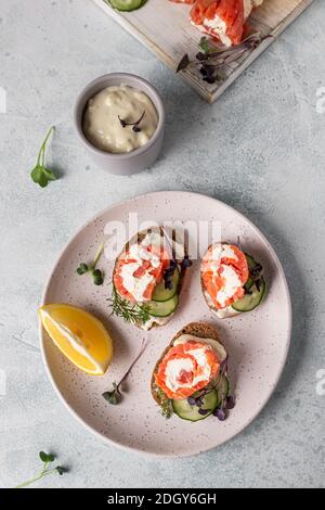Mini-Sandwiches mit geräuchertem Lachs, Frischkäse, Gurke und Microgreen auf Roggenbrot, grauer Steinhintergrund. Vorspeise mit Lachs und Käse c Stockfoto