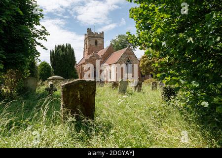 Dunchurch, Warwickshire, UK - 16/06/20: St. Peter's Church in einem teilweise bewachsenen Kirchhof, von dem alte Steingrabsteine in langem Gras stehen. Stockfoto